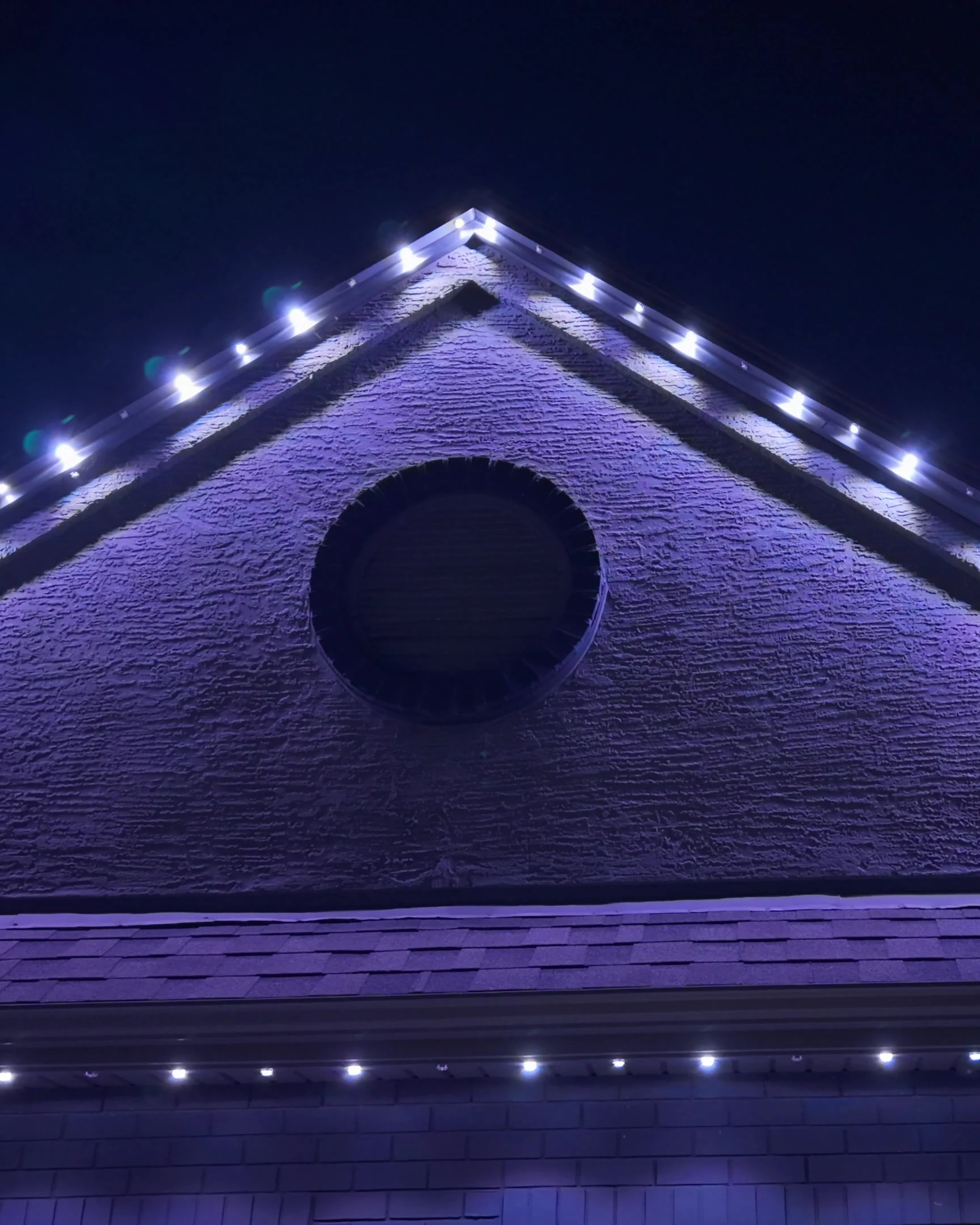 A stucco gable at night, white emitters evenly spaced along track fitted under the eave from peak to corner.