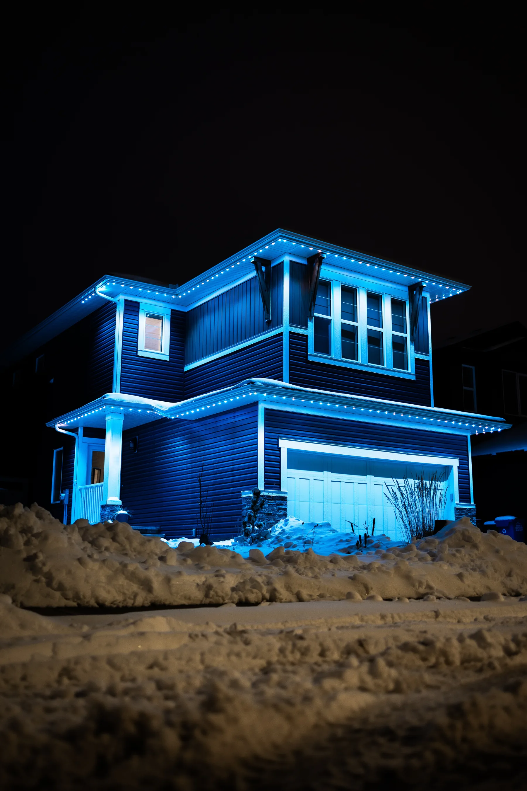 A two-storey Alberta home behind a snowbank at night, every roofline lit in glacier blue.
